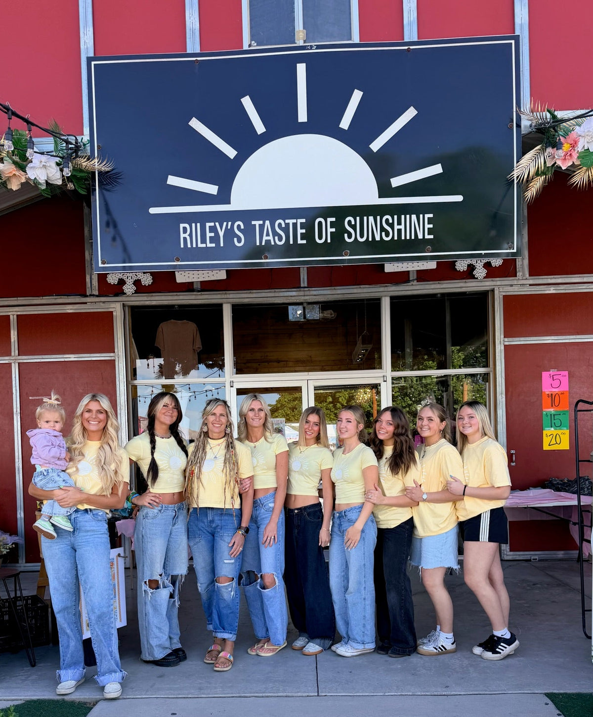 A group of ten young women posing in front of a red building with a large black sign that reads 'RILEY'S TASTE OF SUNSHINE.' They are wearing matching yellow t-shirts and jeans, with one woman holding a baby.